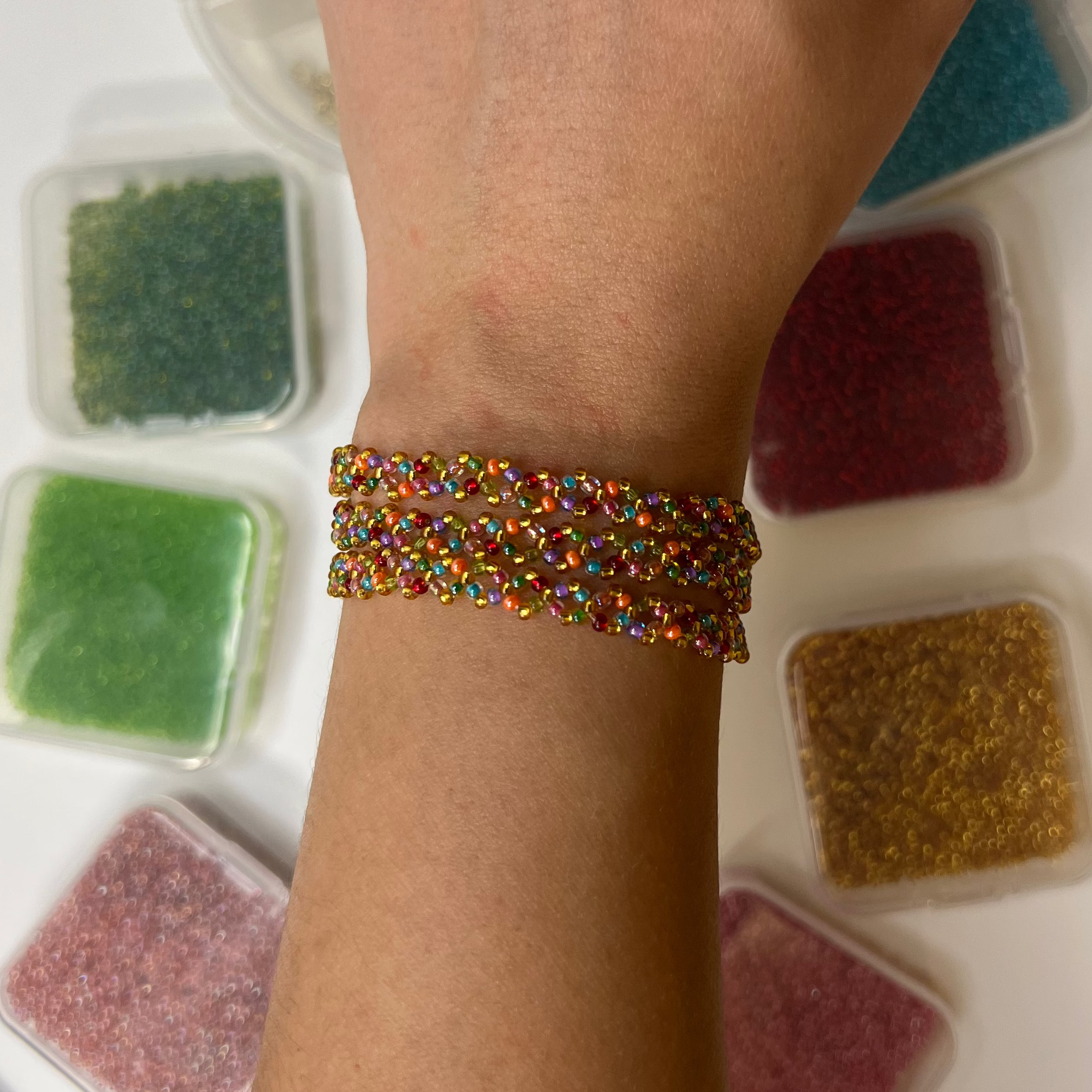 Hand with a colorful bracelet with  small containers of colored beads in the background on a white surface.
