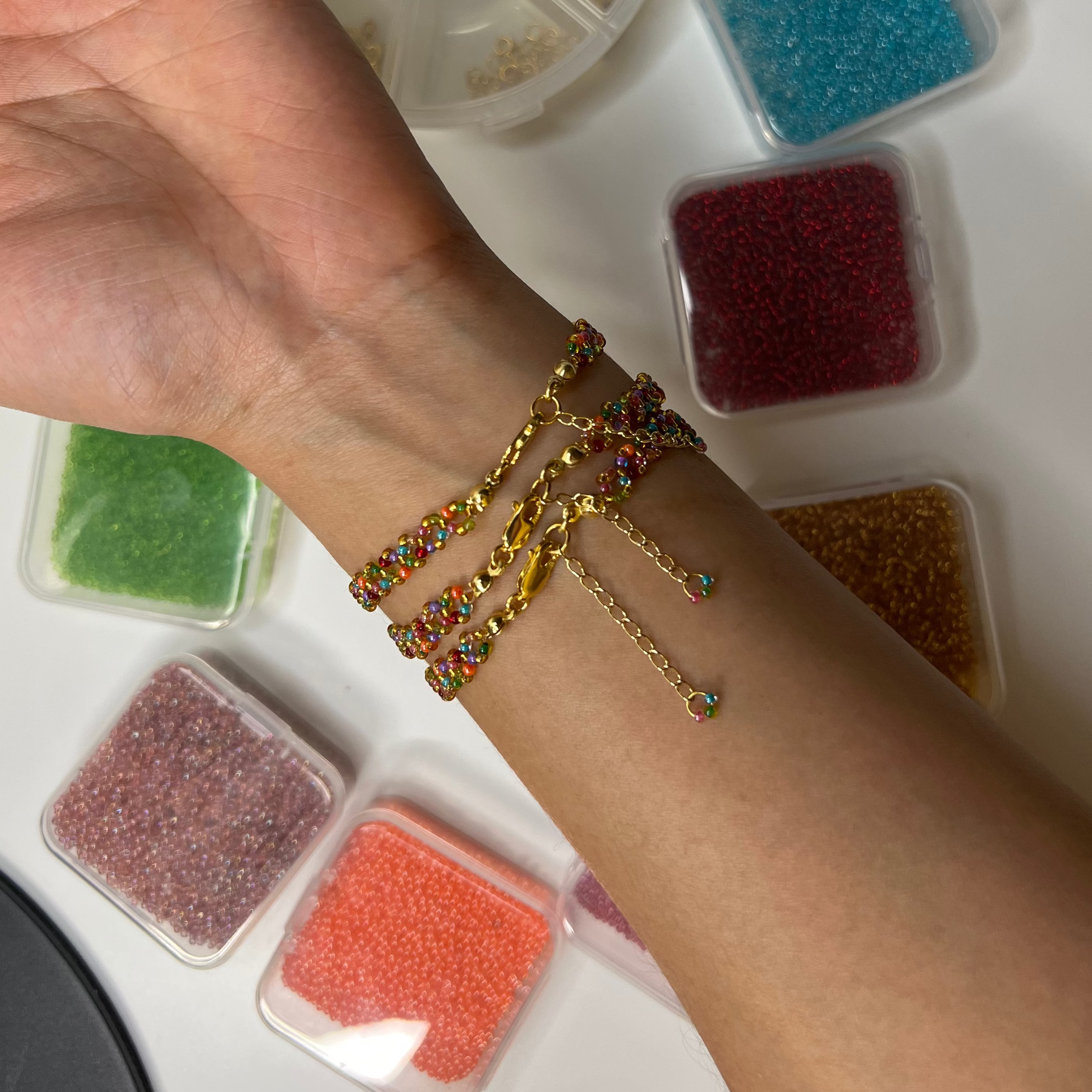 Hand wearing gold bracelets with small containers of colored beads on a white surface