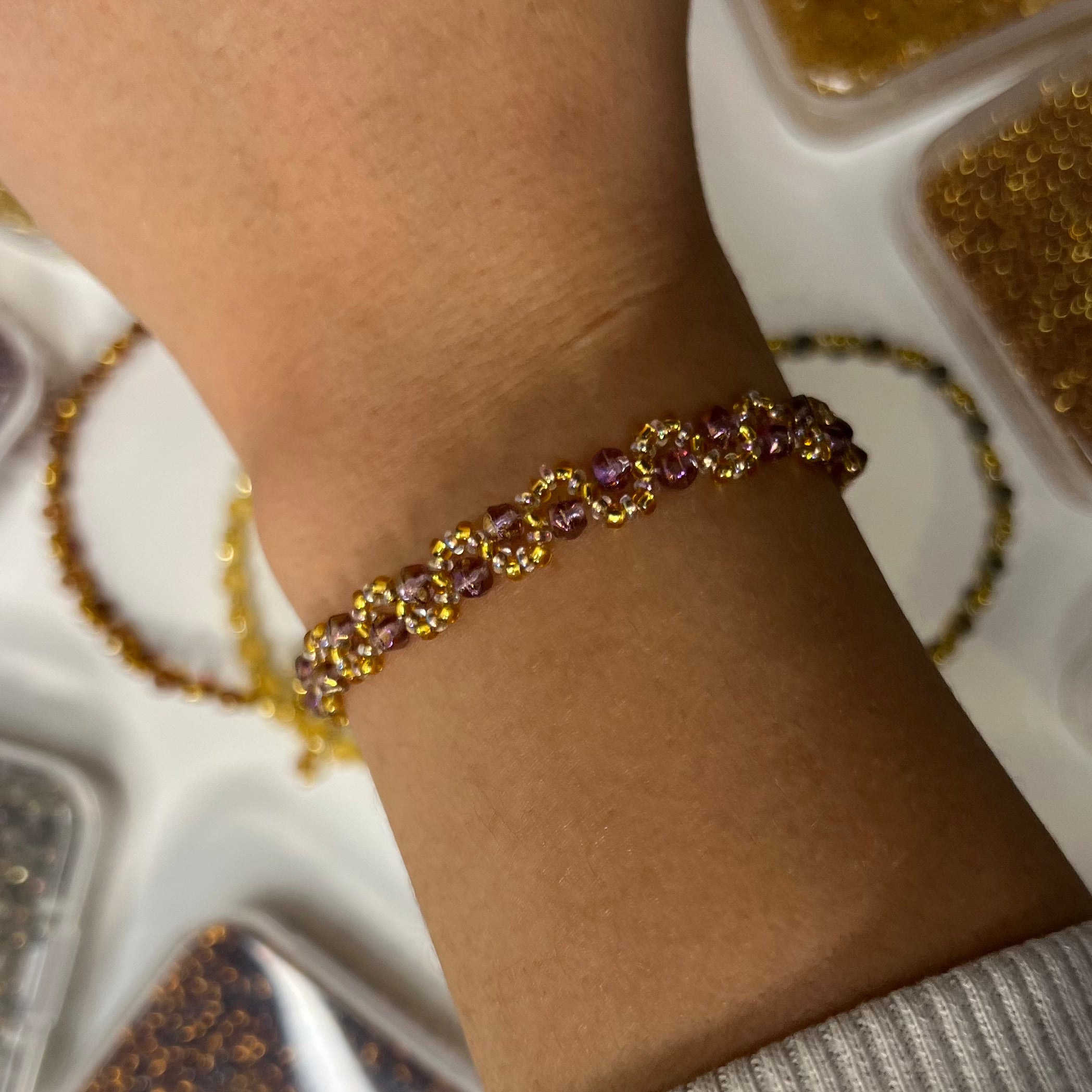 Close-up of a wrist wearing a gold and purple beaded bracelet with a decorative background.