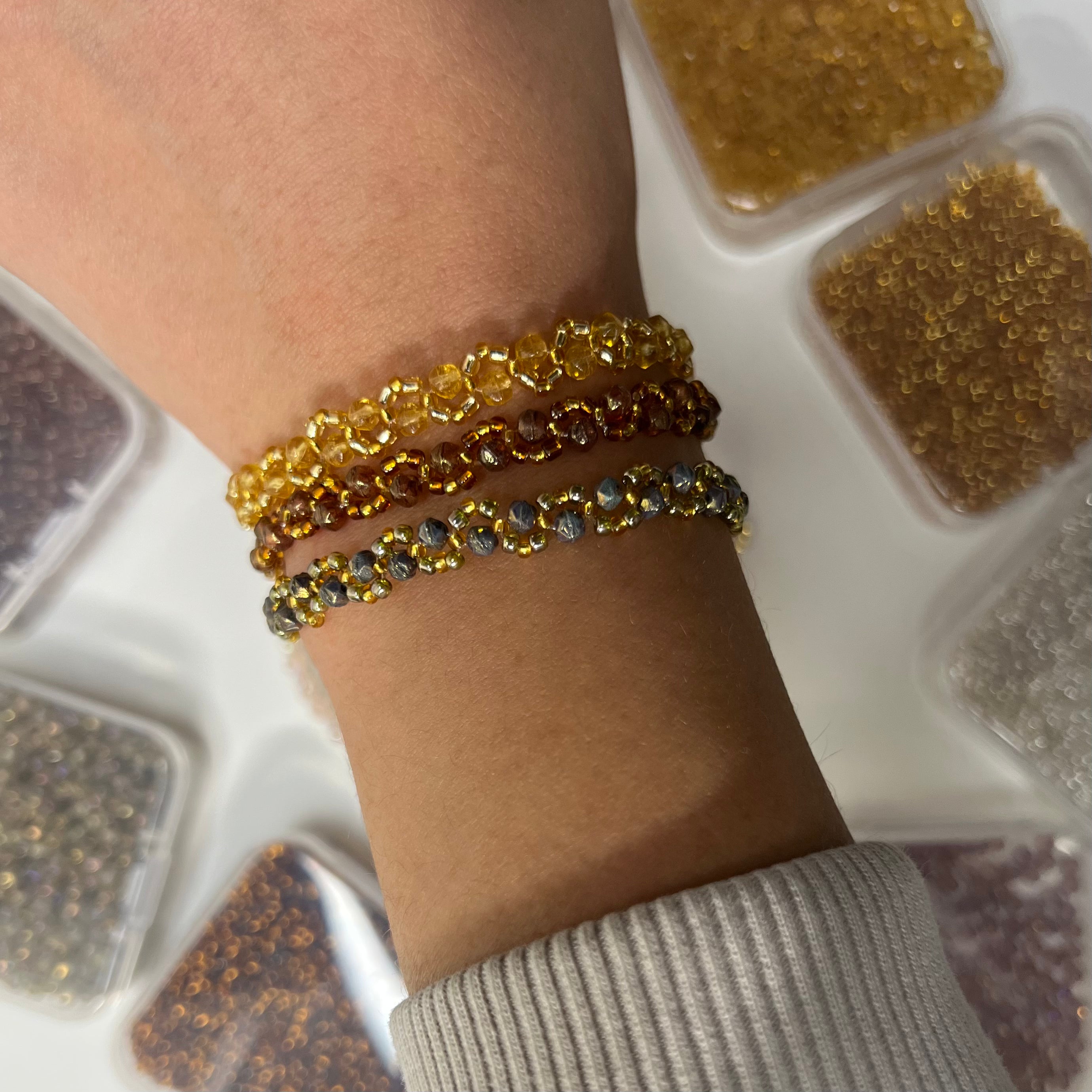Close-up of a wrist wearing three gold bracelets with a container of beads in the background.
