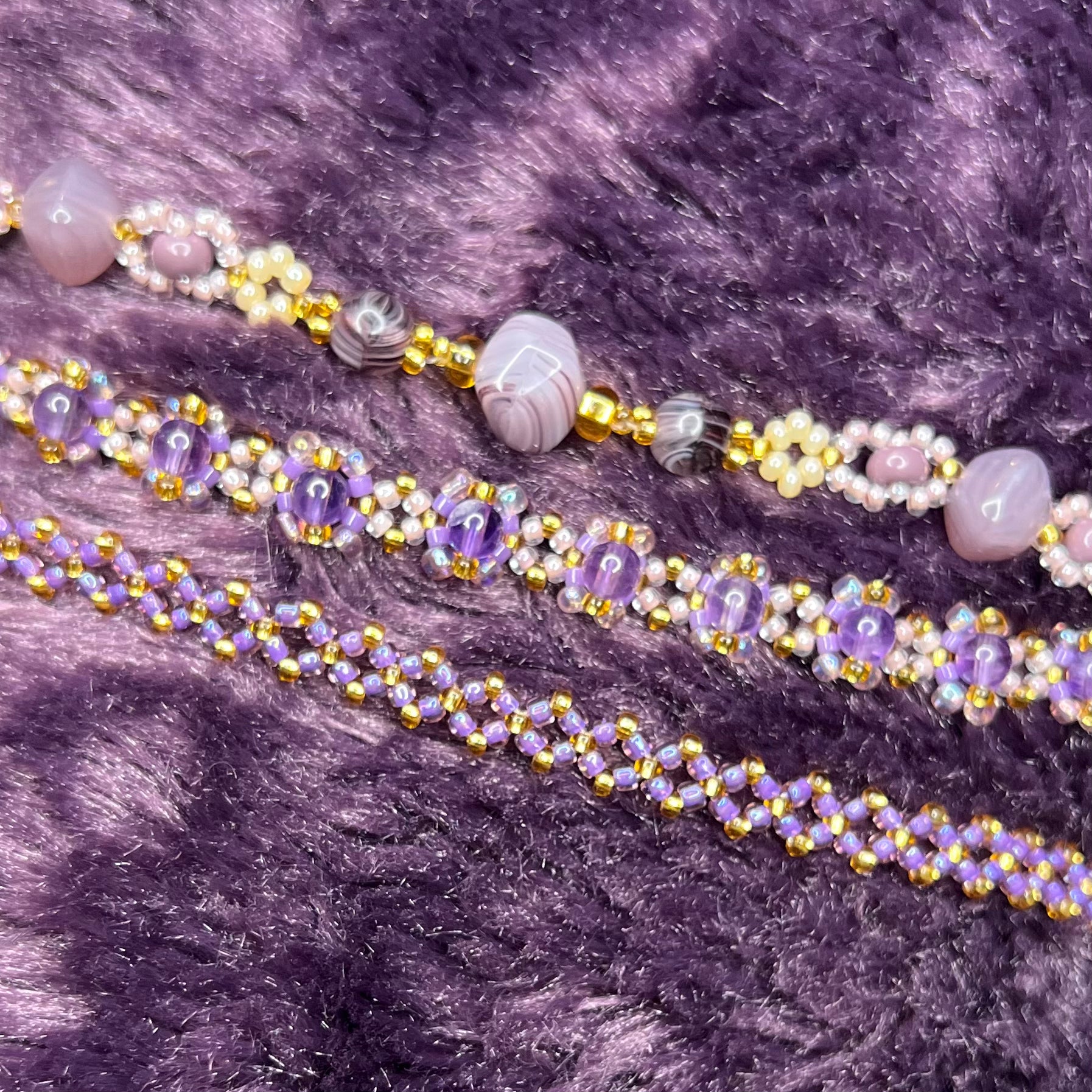 Three beaded bracelets with purple and gold beads on a textured purple fabric background.